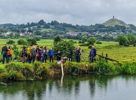 People standing on the edge of a river with Glastonbury Tor in the background.