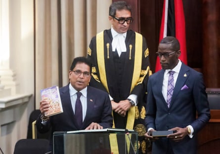 A middle-aged man holds a copy of the Bhagavad Gita as two men, one in speaker’s robes, look on
