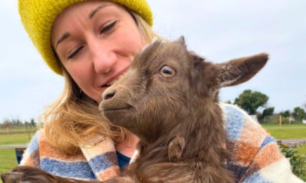 A woman in a plaid coat cuddles a brown baby goat, at Flowerdew Farm, Dorset.
