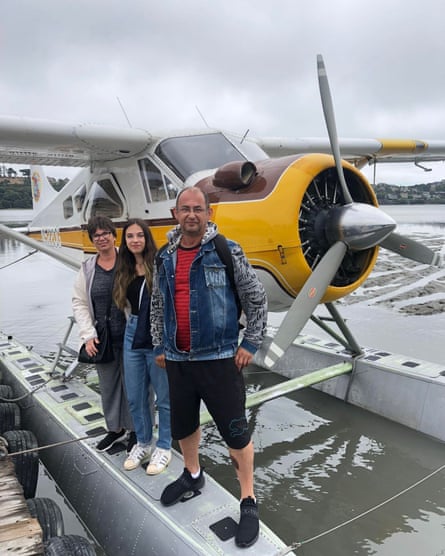 A mother, father and their older daughter stand in front of a seaplane against a cloudy sky.
