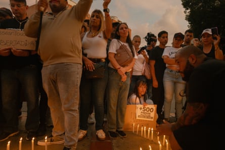 Jeisi Blanco sitting on the ground behind a ring of candles and holding a small sign, surrounded by a crowd of people