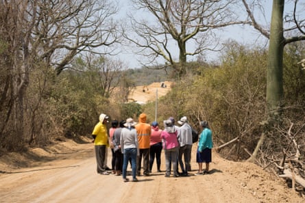 Um grupo de pessoas visto de trás está em uma estrada de terra olhando para uma escavadeira e um caminhão basculante em um terreno limpo à distância