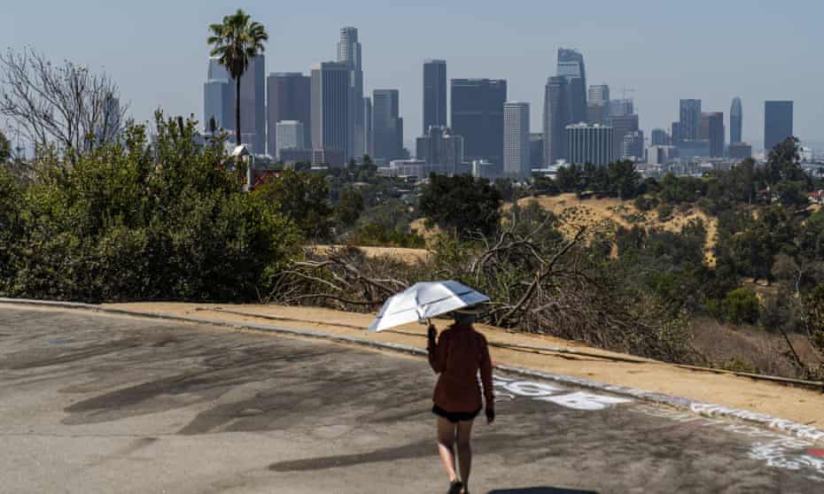 A man walks a park path while holding a sun protection umbrella with the Los Angeles skyline in the distance.