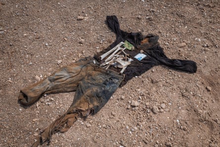 A black long-sleeved shirt and a pair of ragged jeans are laid out on a patch of stony sand, with a couple of bones, an ID card and a bank note.