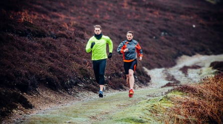 Two fell runners running past heather and foliage across a hilly landscape. Following a dirt track they run down side by side.