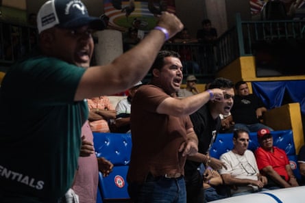 Man pumps his fist in air while another standing has his mouth open and more watch seated in blue chairs
