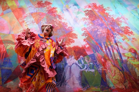 A man in a pink, orange and yellow outfit stands in front of a bright artwork.