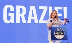 Meloni, smiling, adjusts the microphone at a lectern, while behind her on a backdrop is the word 'Grazie!' in large white letters on an azure backdrop