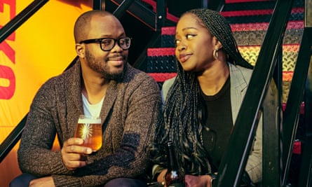 Anthony and Helena Adedipe sit on black metal stairs, they are looking at each other and he is holding a glass of beer and she is holding a bottle of beer