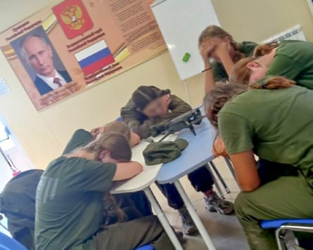 A group of girls in military clothing sit studying at a desk. A poster of Vladimir Putin is on the wall behind them.