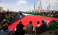 Houthi supporters sit around a huge Palestinian flag in a public space with a mosque in the background