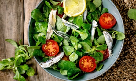 Green field salad with pickled anchovies or sardine fillet, and cherry tomatoes, served in blue bowl with lemon and olive oil on straw napkin over old wooden background.