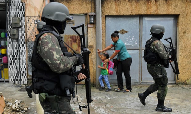 A mother and child walk past military police on patrol near the Vila Kennedy favela in Rio de Janeiro on February 23, 2018.