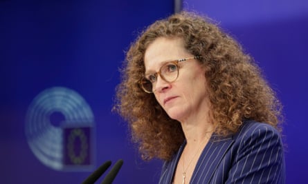 A white woman with long, thick, curly reddish-brown hair wearing round tortoiseshell glasses and a blue pinstripe suits appears to be listening to someone as she stands behind a microphone at a lectern, with a bright blue background behind her.
