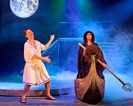 Toby Park in a toga, gesturing and laughing, next to Aitor Basauri in a cloak, standing on a boat with an oar, with mist on the stage and a picture of the moon in The Frogs
