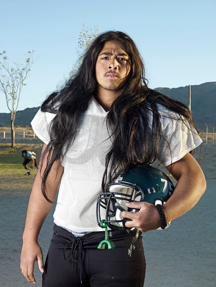 Portrait of a young American football player. He is holding his helmet revealing his very long dark hair hanging over his football shirt