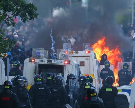 Riot police in helmets stand near white police vehicles with a large fire burning in front them and facing crowds of people