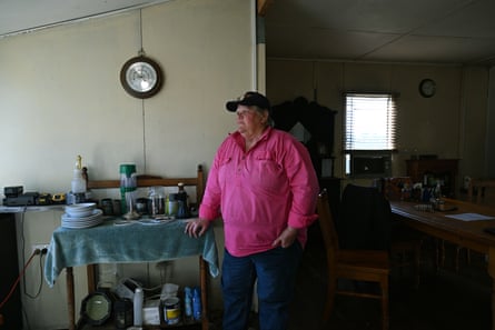 Trish Goodwin wears a baseball cap and bright pink shirt. She stands in her home leaning on a wooden shelf containing crockery, coffee and other paraphernalia