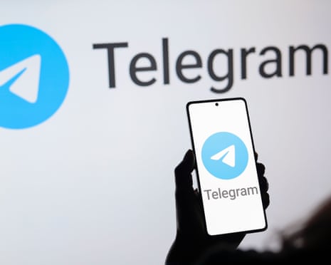 A hand holds a phone displaying the Telegram logo in front of a sign also displaying the Telegram logo