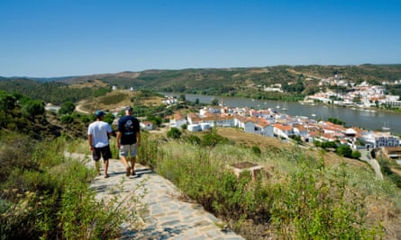 Two men in T-shirts and shorts walk on a path above a river lined with buildings