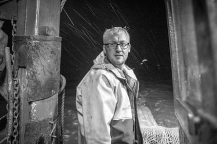 A black and white photo of man wearing a fisher’s waterproof jacket and glasses standing at the back of a trawler heading out to sea