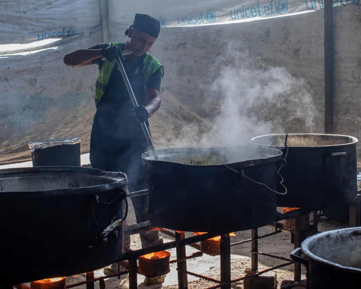 Man stirring a large pot in a community kitchen in a tent