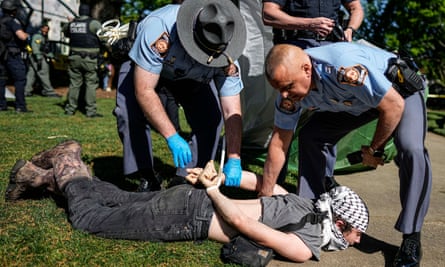 Two officers have a young white man prone and face down on the ground with his wrists zip-tied behind him.