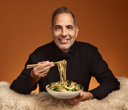 Chef Yotam Ottolenghi leaning on a fleecy chair, feet up, holding a bowl of green tea noodles