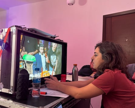 A woman sits in front of a screen that shows another woman made up and dressed as a man
