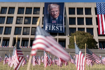 large banner of trump saying ‘american workers first’ on building, with many american flag signs in grass in front
