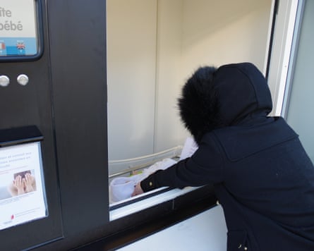 A person in a black coat and hood deposits a doll in a baby hatch