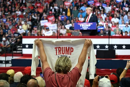 Seen from behind, blond middle-aged woman holds up white sweatshirt with red letters spelling Trump toward Donald Trump on stage.