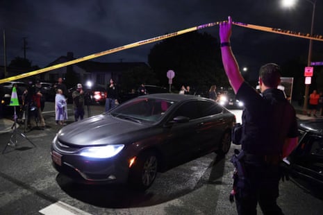 A uniformed officer, lit up by the red and blue lights of emergency vehicles on a dark night, lifts up yellow crime scene tape to allowan unmarked sedan to drive through while the media and other onlookers watch from the other side of the road.