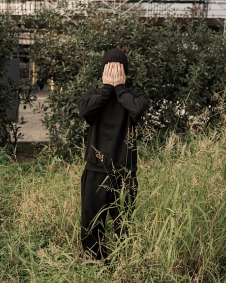 a young man stands in tall grass with his hands over his face; he wears a black woolly hat, baggy sweatshirt and sweatpants.