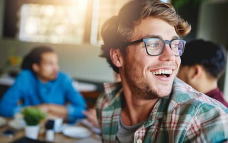 A smiling young man sitting at a cafe table with friends in the background
