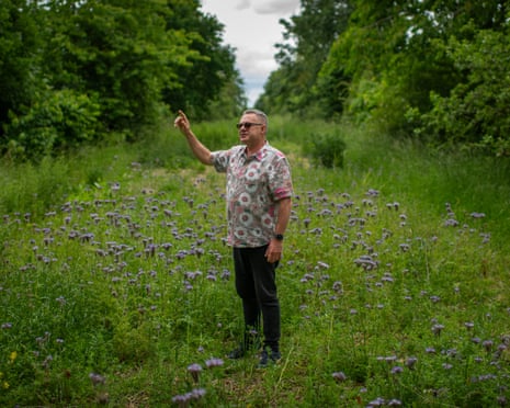 Wolfe stands in a wildflower meadow between rows of trees, gesturing upward with one hand