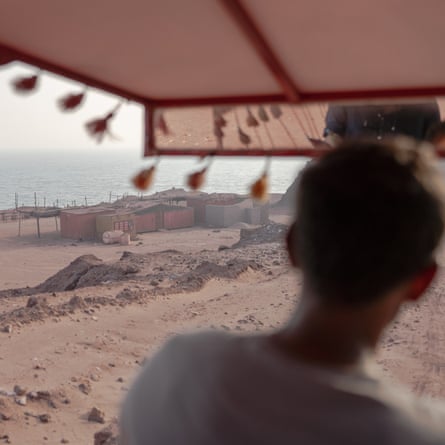 A tour guide looks out from a shaded hut over sandy terrain toward buildings and the sea