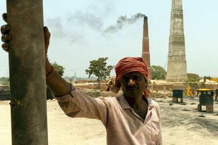 A man with a covered head stands in front of two tall chimneys, one giving off smoke.