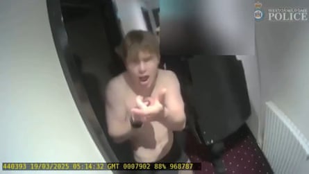 Screengrab of video shows Natland barechested and apparently shouting with his mouth open, holding one hand out in a gun shooting gesture. He is standing in the doorway of a hotel room.