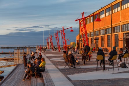 People relax around cafe tables and on steps at a modern harbourfront development in Tallinn, Estonia; there is a restored redbrick industrial building to one side and bright red cranes with suspended lamps along the promenade. It looks cold and wintry.