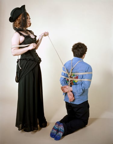 A woman in black Victorian-style dress holds a rope binding a kneeling man’s wrists