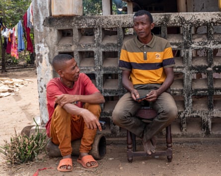 Two black men, one sitting on a log, the other sitting on a chair, in conversation