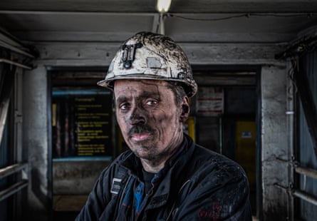 A miner after his shift covered in coal dust looks at the camera. He is wearing overalls and a white helmet.