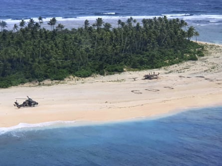 a helicopter sits on a beach near a grove of trees and SOS spelled out on the sand in leaves