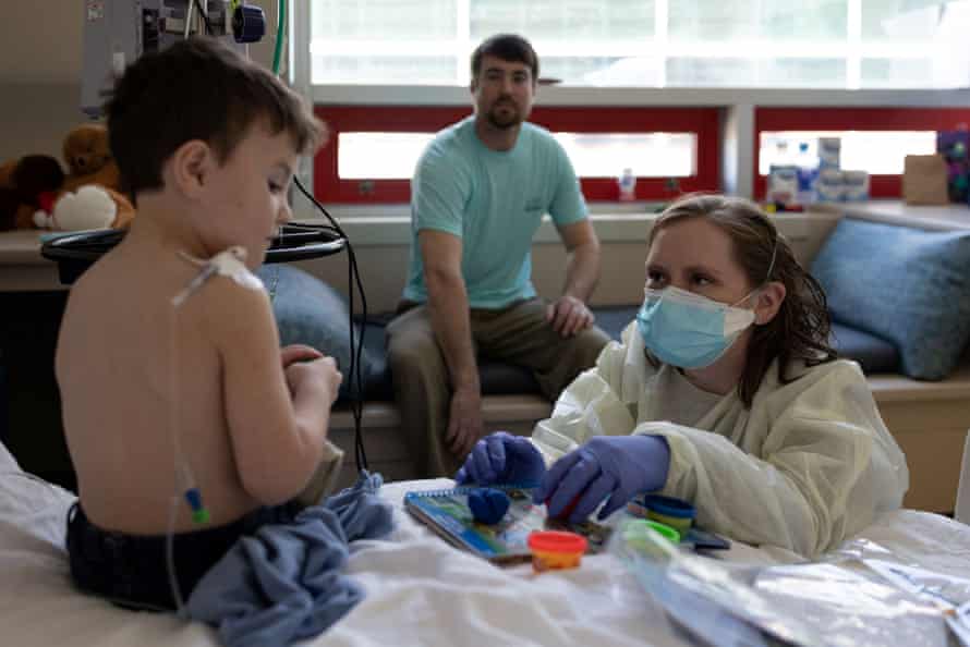 A child sits on a bed in a hospital room playing with Play-Doh with a medical worker who is wearing scrubs, a mask and gloves. A man looks on in the background.