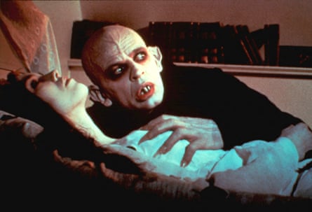 A woman lying down in an open grave, and a man with a ghostly white face clutching her chest