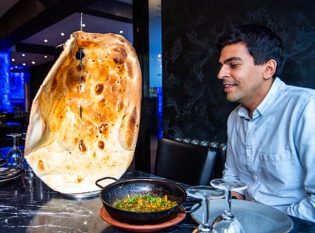 A man sat a table in a restaurant with a bowl of curry and a giant naan skewered on a tabletop plinth