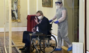 man in a wheelchair and face mask waves, an attendant in heavy protective wear behind him