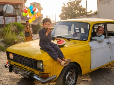 a boy sits on the bonnet of a stationary battered yellow car while an elderly man leans his elbow out of the driver's side window