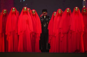 At the centre is The Weeknd dressed in black, adorned with glittering embellishments, holding a microphone. He is flanked by individuals wearing red robes and golden face masks as part of his show in Vancouver, Canada.